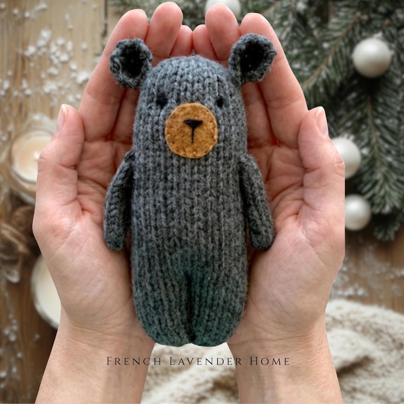 May include: A small, hand-knitted dark gray bear toy with a round, tan-colored felt muzzle, black embroidered nose and mouth, and two small, rounded ears. The bear has a textured, ribbed knit pattern throughout its body and limbs.