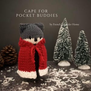 May include: A knitted fox toy wearing a red cape with white trim. The fox has gray ears and a white face. The text "CAPE FOR POCKET BUDDIES" is above the fox, and "by French Lavender Home" is below.