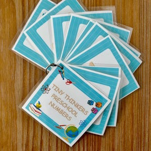 May include: A set of flashcards for preschool children, featuring a teal and white design. The cards are held together by a metal ring and include the text "Tiny Thinkers Preschool Numbers" along with educational illustrations.