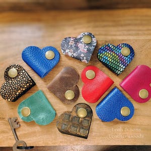 May include: A collection of ten heart-shaped leather cord organizers, each featuring a brass-colored metal snap button. The organizers come in various colors and textures, including blue, red, pink, green, brown, floral, and iridescent scale patterns.