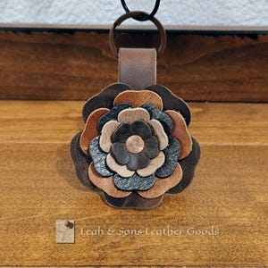 May include: A leather keychain featuring a multi-layered flower design in shades of dark brown, tan, and black. The flower petals are stacked and secured with a central copper-colored rivet. A brown leather loop attaches the flower to a metal ring.