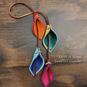 May include: A leather accessory featuring four colorful, leaf-shaped pendants in red, orange, yellow, teal, blue, and pink. Each pendant is attached to a brown leather cord, with a loop at the top. The text "Leah & Sons Leather Goods" is visible.