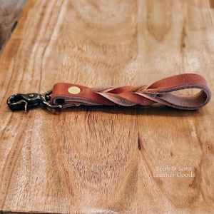 May include: A tan leather keychain featuring a braided strap design, a single gold-tone metal rivet, and a dark metal swivel lobster clasp. The text Leah & Sons Leather Goods is visible in the lower right corner of the image.