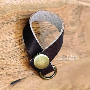 May include: A dark brown leather loop strap featuring a circular brass-toned metal snap button closure and an attached brass-toned metal ring. The text Leah & Sons Leather Goods is printed in white on the right side of the leather strap.