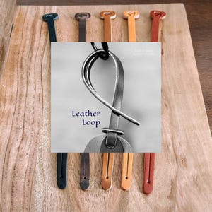May include: A collection of leather loops in various colors including black, dark brown, tan, and reddish-brown. The central focus is a dark gray leather loop with a knotted design, featuring the text Leather Loop and Leather Goods.