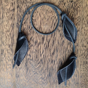 May include: A black leather necklace featuring a thin cord and three distinct leaf-shaped pendants. Each pendant is constructed from folded leather pieces with a smooth outer surface and a textured inner side, attached to the cord with small loops.