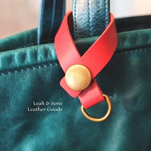 May include: A red leather strap with a brass-toned snap button and a circular ring attached to a dark green leather bag. The text Leah & Sons Leather Goods is printed in white on the surface of the dark green leather bag.