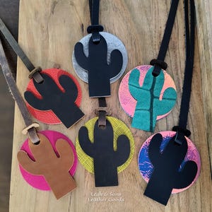 May include: Six leather cactus-shaped ornaments with circular backings in various colors and textures, including red, silver, yellow, and patterned designs. Each ornament features a black or brown leather strap and the text Leah & Sons Leather Goods.