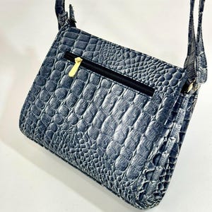 May include: A blue crossbody bag with a faux crocodile skin texture. The bag has a black zipper with a gold-colored pull and a matching strap. The bag is angled, showing the front and side.