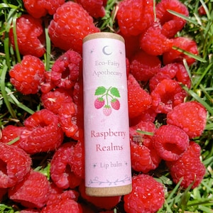 May include: A cylindrical lip balm tube with a light pink label featuring a crescent moon icon, the text Eco-Fairy Apothecary, a graphic of three raspberries, and the text Raspberry Realms Lip balm, surrounded by a cluster of fresh red raspberries.