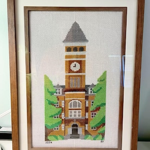 Peut inclure: Tableau de broderie encadré représentant un bâtiment universitaire avec une tour d'horloge. L'œuvre utilise des tons d'orange, de marron et de vert, sur fond blanc. Le cadre est en bois foncé. L'année "2024" est visible.