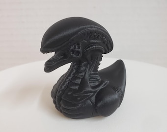May include: A black 3D printed rubber ducky shaped like an alien creature. The ducky has a detailed head and body with a textured surface.
