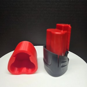 May include: A red and black power tool battery and its protective cap. The battery is red with a black base. The cap is red and has a unique shape. The items are on a white surface against a dark background.
