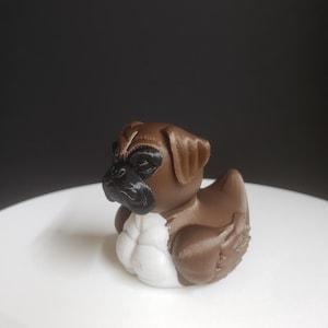 Könnte beinhalten: Eine braune Gummiente mit einem Mopsgesicht und weißen Brustmuskeln. Die Ente sitzt auf einer weißen Oberfläche.