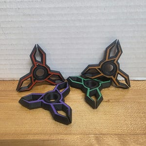 Puede incluir: Cuatro spinners de colores en rojo, marrón, verde y morado. Cada spinner tiene un diseño de tres puntas con un rodamiento central. Los spinners están dispuestos sobre una superficie de madera.