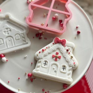 Puede incluir: Una galleta blanca con forma de casa de jengibre con chispas rojas y doradas, un lazo rojo y un cortador de galletas rosa en forma de casa, están sobre un plato blanco. Otras galletas con temática navideña son visibles sobre una superficie roja.