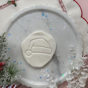Może przedstawiać: Biała wykrawaczka do ciastek w kształcie czapki Świętego Mikołaja na talerzu z marmurowym wzorem. Talerz otoczony jest elementami dekoracyjnymi, w tym białym, falistym brzegiem, konfetti w kształcie płatków śniegu i świąteczną zielenią. Tło jest w kolorze jasnoróżowym.