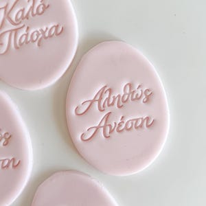 Puede incluir: Decoraciones rosas pálidas en forma de huevo con escritura griega. Las decoraciones están sobre una superficie blanca. El texto incluye frases como "Felices Pascuas" y "Muchos años".