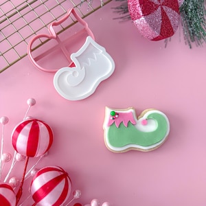 Puede incluir: Imagen navideña rosa con un cortador de galletas y una galleta decorada con forma de zapato de elfo. La galleta es verde, rosa y blanca. También hay adornos y ramitas verdes.