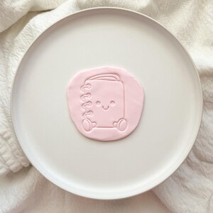 May include: A light pink circular piece of clay featuring an embossed design of a spiral-bound notebook with a smiling face, two small circular eyes, and two rounded feet at the bottom, centred on a smooth white plate.
