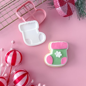 Puede incluir: Un cortador de galletas rosa con forma de calcetín navideño y un corazón. Una galleta de calcetín navideño decorada con glaseado verde, rosa y blanco. Adornos decorativos a rayas rojas y blancas y bayas rosas.