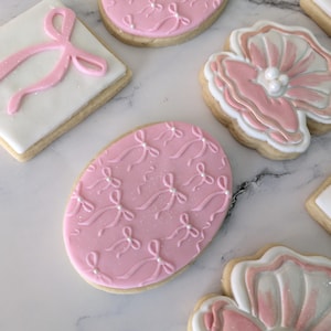 Puede incluir: Surtido de galletas de azúcar decoradas en rosa y blanco. Las galletas están decoradas con lazos, perlas y diseños florales.