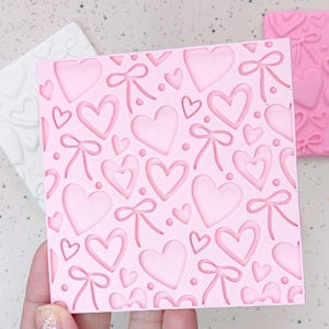 May include: Pink silicone mold with a repeating pattern of hearts and bows. The mold is designed for baking and crafting.