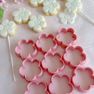 May include: Pink flower-shaped cookie cutters and flower-shaped cookies on a pink surface. The cookies have white icing and are on sticks. Decorative pink flowers are in the background. The cookie cutters are made of plastic.