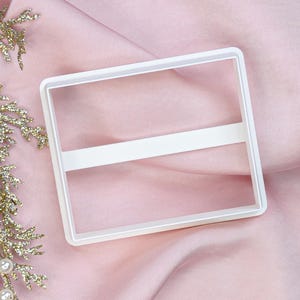 May include: White plastic rectangle cookie cutter with a horizontal line across the middle.