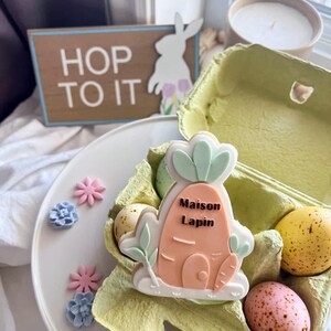 Könnte beinhalten: Eine Osterdekoration mit einem karottenförmigen Keks mit der Aufschrift "Maison Lapin" in einem Eierkarton mit dekorierten Eiern. Ein Schild mit der Aufschrift "Hop to it" ist zu sehen, zusammen mit dekorativen Blumen.