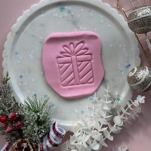 Puede incluir: Un molde de arcilla rosa con un diseño de caja de regalo está centrado en una bandeja de mármol blanco. La bandeja está decorada con confeti de copos de nieve. Adornos navideños y vegetación rodean la bandeja.