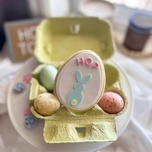 Puede incluir: Una composición temática de Pascua con una galleta con forma de huevo decorada, con la palabra "HO" y un diseño de conejito. Está en una huevera amarilla con huevos moteados. El conjunto está sobre un plato blanco.