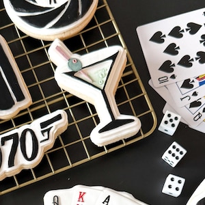 May include: Assorted decorated sugar cookies with a spy theme. The cookies include a martini, playing cards, dice, and a gun. The cookies are white with black and red accents, arranged on a gold wire rack and black surface.