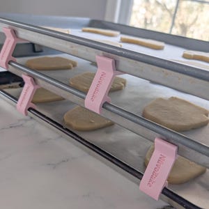 Puede incluir: Dos bandejas para hornear con pinzas rosas que dicen "Now Baking" están apiladas una encima de la otra. Las bandejas están forradas con papel para hornear y contienen varias galletas en forma de corazón.