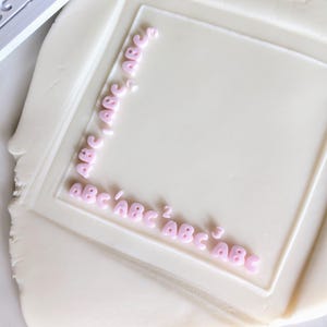 Può includere: Un tagliapasta quadrato con le lettere "ABC" rosa, premuto in un impasto bianco. Le lettere sono disposte a forma di L all'interno del quadrato. Un tagliapasta simile separato è visibile in alto a sinistra.