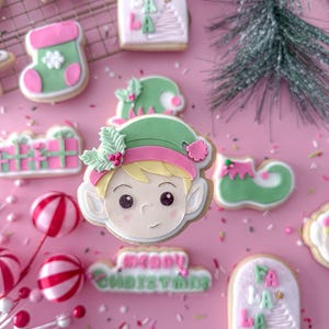 May include: A collection of festive Christmas cookies, including an elf face, a stocking, and a gift box, arranged on a pink surface. The cookies are decorated with pink, green, and white icing. Some cookies have the text "Merry Christmas" and "Fa La La".