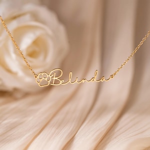 Può includere: Una collana dorata con il nome "Belinda" in corsivo. Un disegno a zampa di cane è posto a sinistra del nome. La collana è presentata su uno sfondo morbido e sfocato.