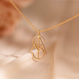 Op de afbeelding: Een goudkleurige hangerketting met een geometrisch ontwerp. De hanger heeft de letters "SA" in een gestileerd, open lettertype, hangend aan een delicate ketting. De ketting wordt gepresenteerd tegen een zachte, vage achtergrond.