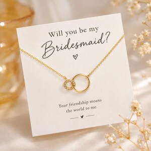 Puede incluir: Un collar dorado con dos círculos entrelazados, uno liso y otro con pequeñas piedras transparentes, sobre una tarjeta blanca con el texto Will you be my Bridesmaid? y Your friendship means the world to me.