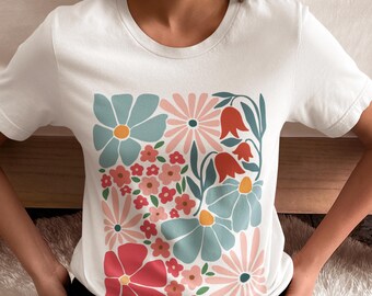 May include: White t-shirt featuring a colorful floral design. The design includes large blue flowers with yellow centers, pink daisies, and red bell-shaped flowers. The shirt is a crew neck style.