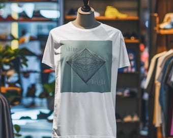 Può includere: T-shirt bianca con un design geometrico a forma di diamante e le parole "shine bright". Il design ha uno sfondo verde acqua. La maglietta è esposta su un manichino in un ambiente commerciale.