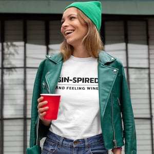 May include: A white t-shirt with the text "GIN-SPIRED AND FEELING WINE" is worn with a green beanie and a green leather jacket. A red plastic cup is held in the hand.