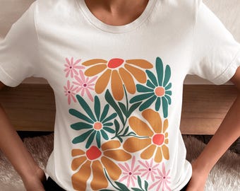 May include: White t-shirt featuring a floral design. The design includes large orange flowers with red centers, teal flowers, and pink flowers. The shirt has a crew neck and short sleeves.