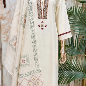 May include: An off-white long-sleeved tunic and matching trousers set featuring a decorative scarf. The tunic has a V-neckline with multi-colored embroidery and small mirror accents. The scarf displays a repeating geometric and floral print.