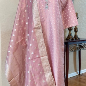 May include: A light pink long-sleeved tunic and matching dupatta set. The tunic features a mandarin collar with floral embroidery along the placket. Both pieces are adorned with a subtle gold polka dot pattern and a metallic gold border.