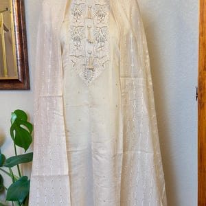 May include: Cream-colored embroidered tunic and matching sheer scarf. The tunic features intricate floral embroidery around the neckline and small, scattered embellishments. The scarf has a delicate, dotted pattern.