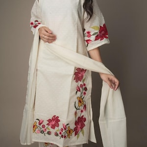 May include: Off-white embroidered kurta set. The ensemble includes a top, pants, and a scarf. The top and pants are adorned with floral embroidery in shades of red, pink, and yellow. The outfit has a relaxed fit and is suitable for casual wear.