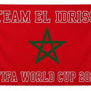 May include: A rectangular red flag featuring a central green five-pointed star. The text "TEAM EL IDRISSI" is printed in white capital letters at the top, and "FIFA WORLD CUP 2026" is printed in white capital letters at the bottom. Metal grommets are visible.