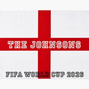 May include: A white flag with a red cross design, bearing the text "THE JOHNSONS" in white. Beneath, the words "FIFA WORLD CUP 2026" are printed in black. White ties are attached at the top corners of the flag.