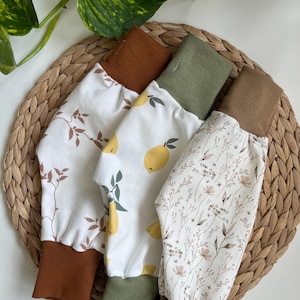 Peut inclure: Quatre paires de pantalons pour bébés avec différents motifs. L'une présente un imprimé citron, une autre un motif floral, et les autres un motif de feuilles marron. Chaque paire a un poignet contrastant marron ou vert.